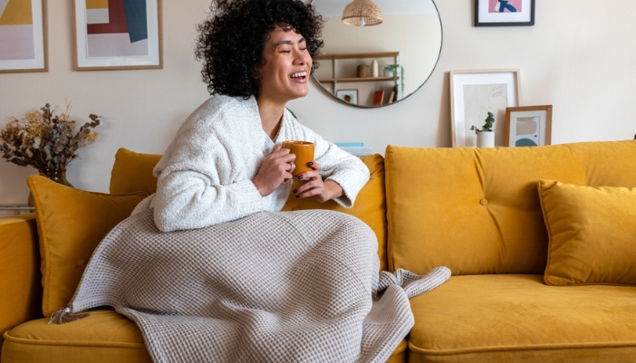 A woman sits on a yellow couch with a gray blanket and a warm cup of coffee. 