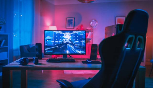 front view of a desk with a monitor, PC, keyboard, and headphones in a modern game room with dim pink and blue LED lights