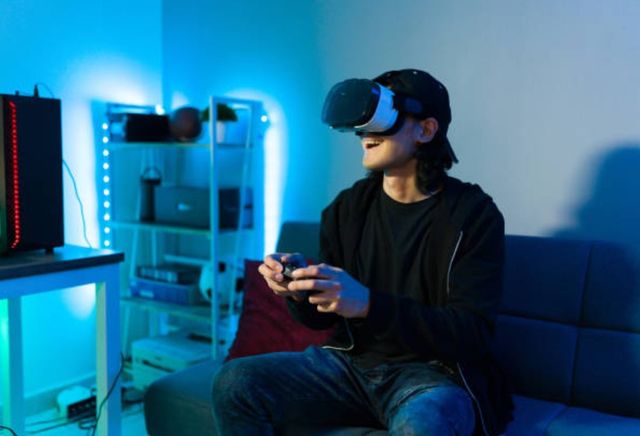 view of a person sitting on a sofa earing a VR headset and holding a controller, with a shelf and deskk with PC and LED blue lights in the background
