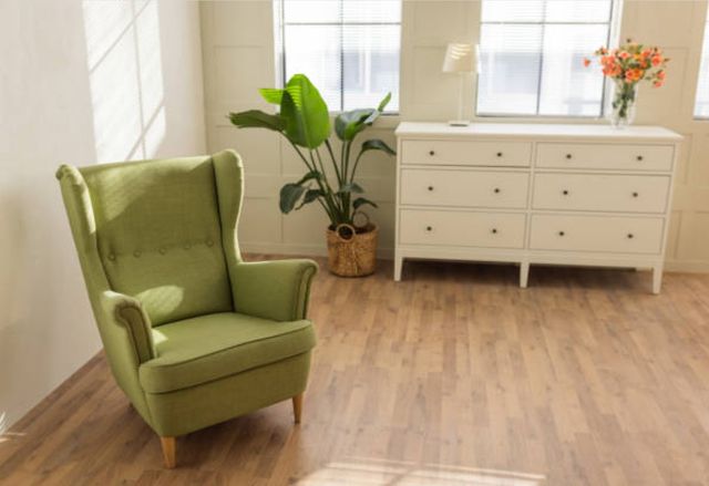 front view of a beige room with wood floors, a white sideboard, and a plant, with a green accent chair in the foreground on the left