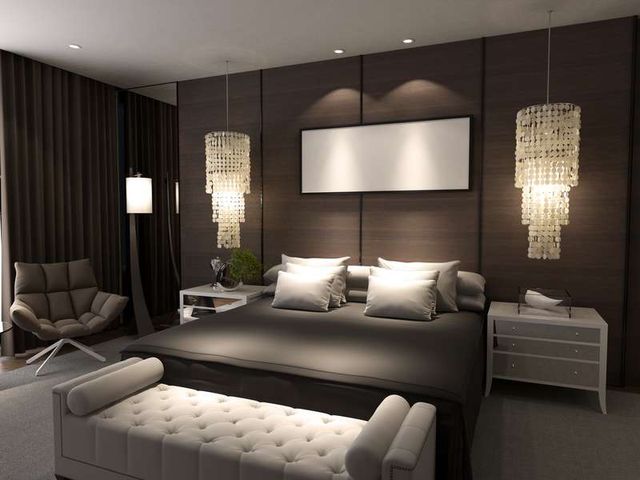 front view of a sleek luxury bedroom with brown panel walls, a large bed, chair, bench, nightstand, and two chandeliers