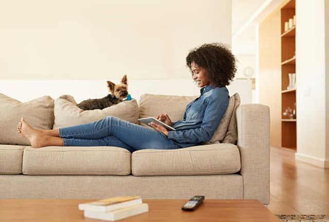front view of a woman sitting with her legs extended on a beige sofa, and a small dog sitting on top of the headrest