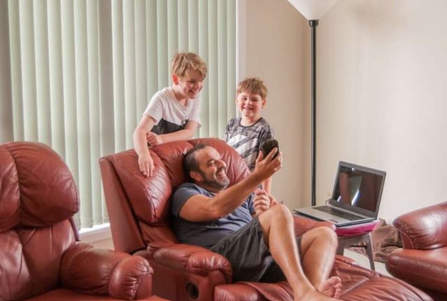 front view of a man sitting in a recliner with two young children around him, as he shows them something on his phone
