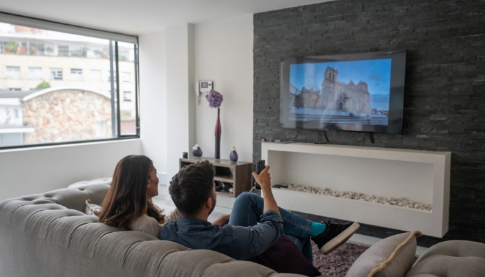 A couple watches tv in their living room with a rock/fireplace entertainment center.