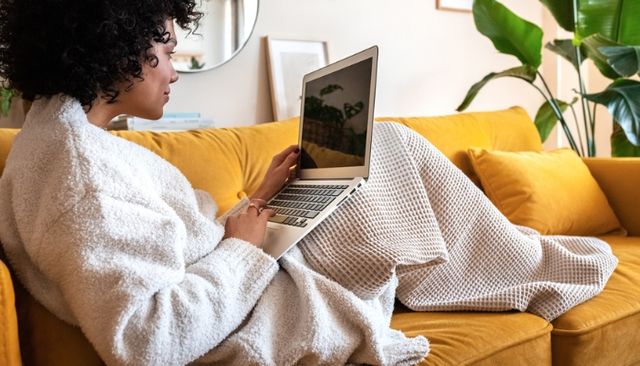 View of a woman sitting and smiling on a yellow sofa while browsing on her laptop