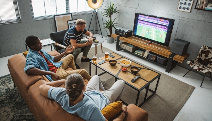 Friends sit together in a living room and watch sports on TV.