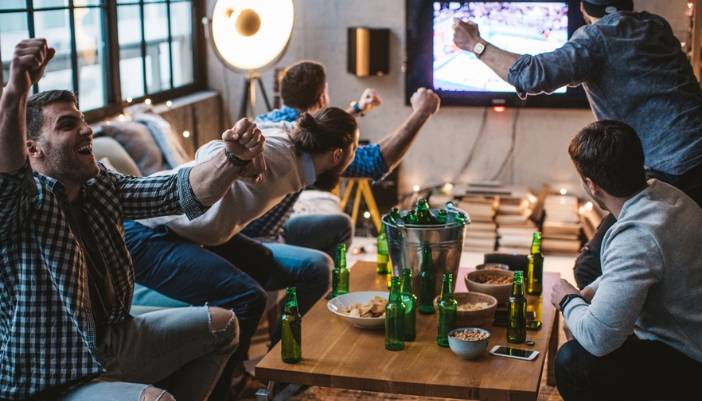 A group of men at home drink, eat, and cheer on sports on the TV.