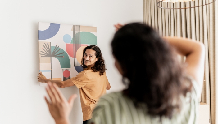 A man and woman hang a colorful piece of art on the wall. 