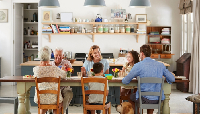 Three generations of a family sit at a large rectangular dining table enjoying a meal. 