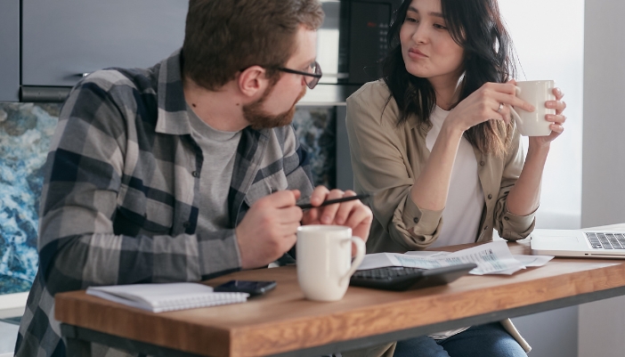 A man and a woman sit at a table drinking coffee with a calculator, laptop, and many papers in front of them.