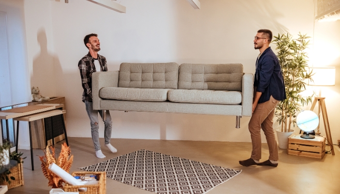 Two men lifting a gray couch out of a minimally decorated living room.