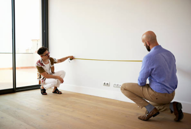 front view of two men kneeling and measuring a white living room wall 