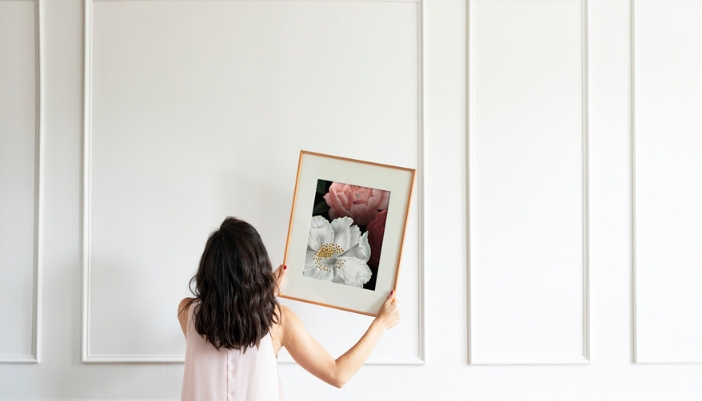 A woman hangs a floral wall art on a white wall space. 
