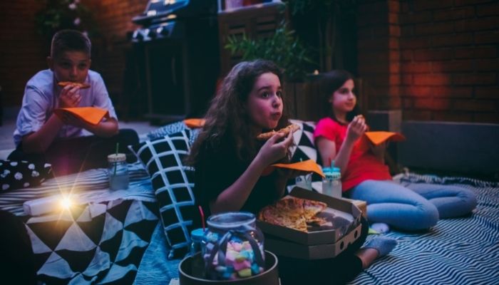 Three kids eating pizza and watching a movie outside