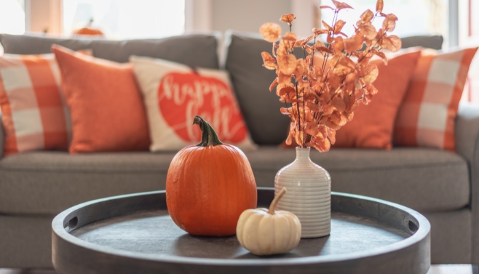A pumpkin and oragne leaves on a coffee table with a gray couch and orange throw pillows behind.
