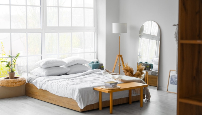 A bright, white bedroom has plants and wooden accents and a tall mirror