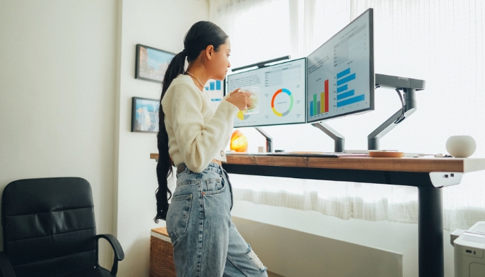 A woman uses a standing desk with multiple monitors and drinking coffee.