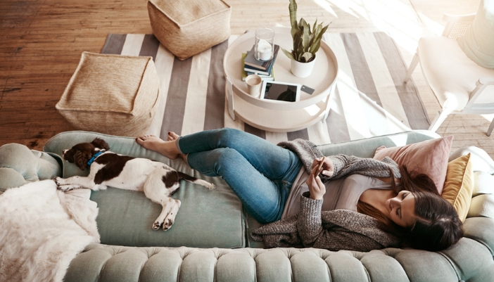 A woman types on her phone while laying with her brown and white dog on a green couch.