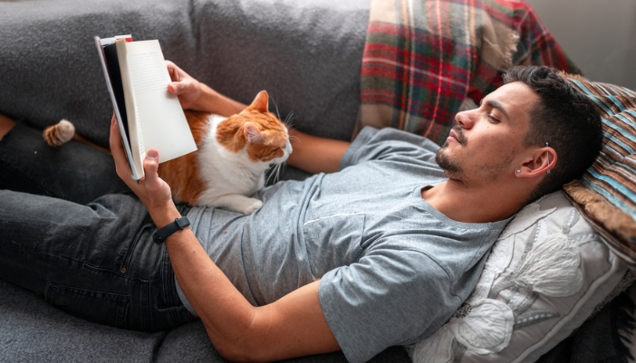 A cat rests on a man’s chest as he lays on his couch and reads a book.