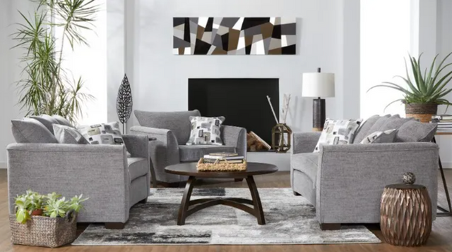 front view of a modern living room with 2 oversized chairs and a loveseat . The furniture is grey with white and grey accent pillows, and the walls are white. The room is decorated with various black, white, grey, and beige decor with plants around.
