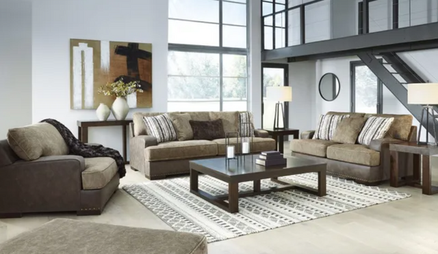 front view of a family room with a fabric sofa, loveseat, and oversized chair. The room has white walls and brown tables and decor.