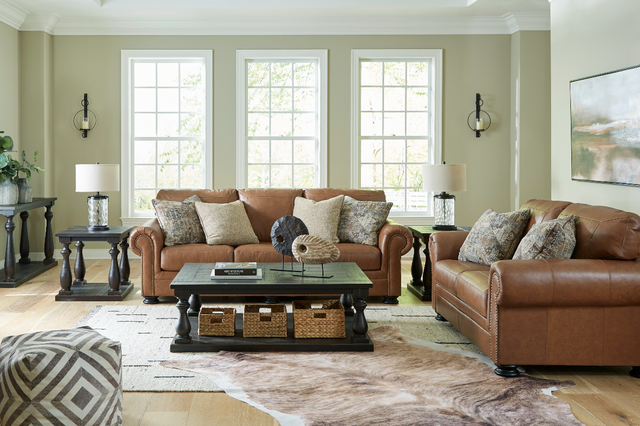 front view of a modern living room with a caramel colored leather sofa and matching loveseat, a black coffee table, and three large windows in the background. The room is decorated with various black, white, and wicker decor.