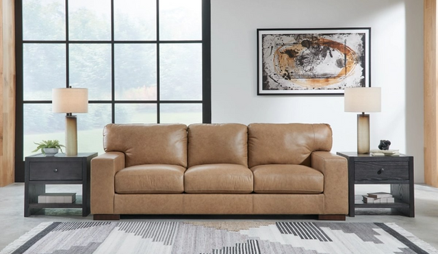 front view of a light tan leather sofa with a black end table on both sides in a contemporary room with a large industrial window
