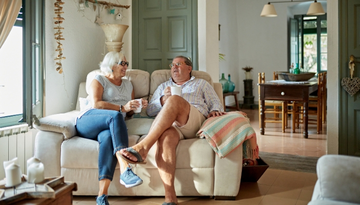 An elderly couple sits together on a white leather loveseat and enjoys a beverage together.
