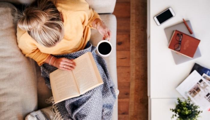 Woman laying back on a tan couch reading a book and drinking coffee
