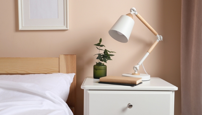 A white desk lamp, plant, and book sits on a white nightstand.