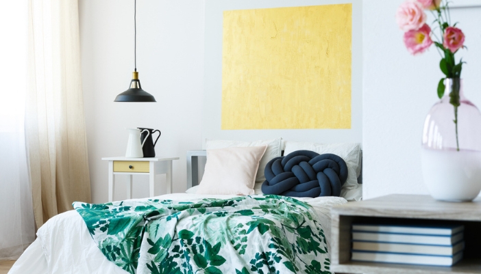 A guest bedroom with white bedding and a green floral throw blanket sits in front of a white wall.