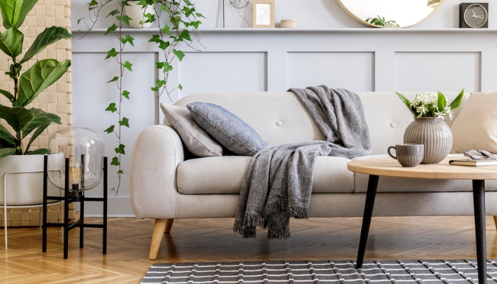 A tan couch sits next to several house plants and with a cozy blanket over it.