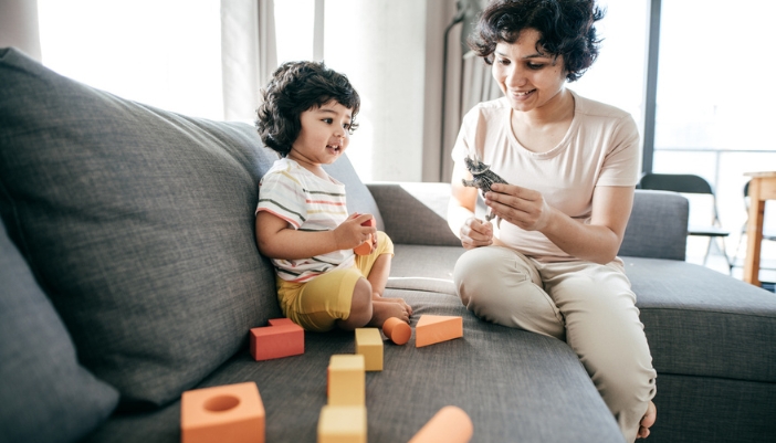 A mom and child sit on a gray sectional and play with toy blocks.