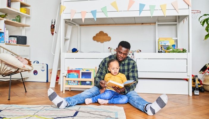 A father and son read a book together in front of a white bunk bed.