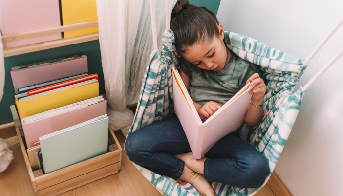A young girl sits in a hammock chair at home reading a book.