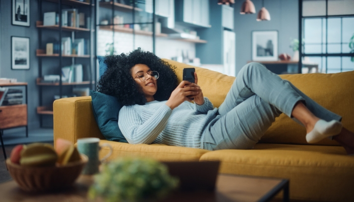 A woman lays on her couch at home and scrolls on her phone.