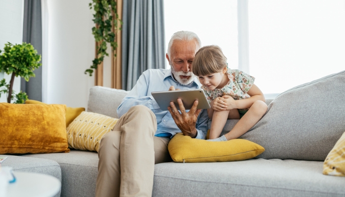 An elderly man sits with a young girl and looks at a tablet together.