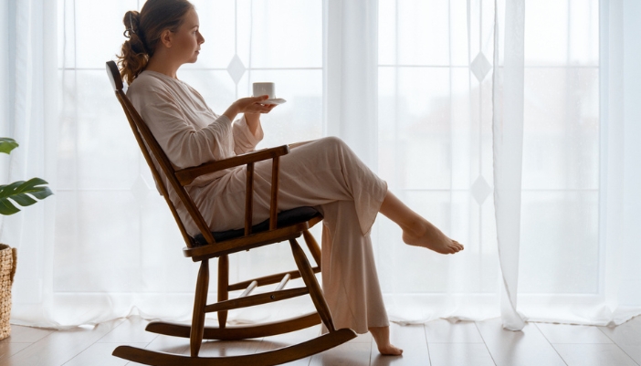 A woman sits in a rocking chair in front of her window and drinks tea.