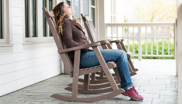 A young woman sits on her porch in a brown rocking chair.