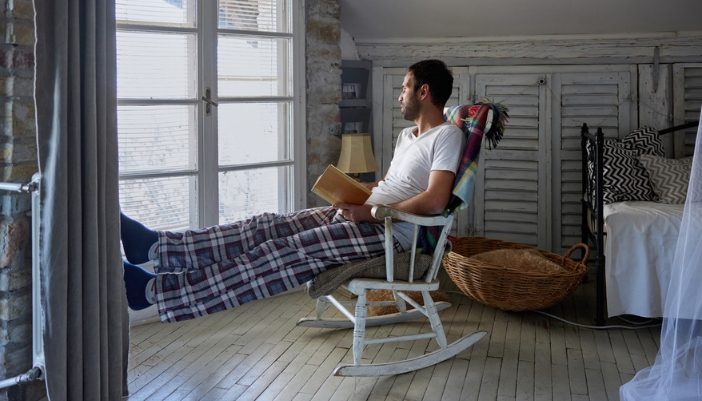 A man sits in his bedroom on a rustic white rocking chair and reads a book.