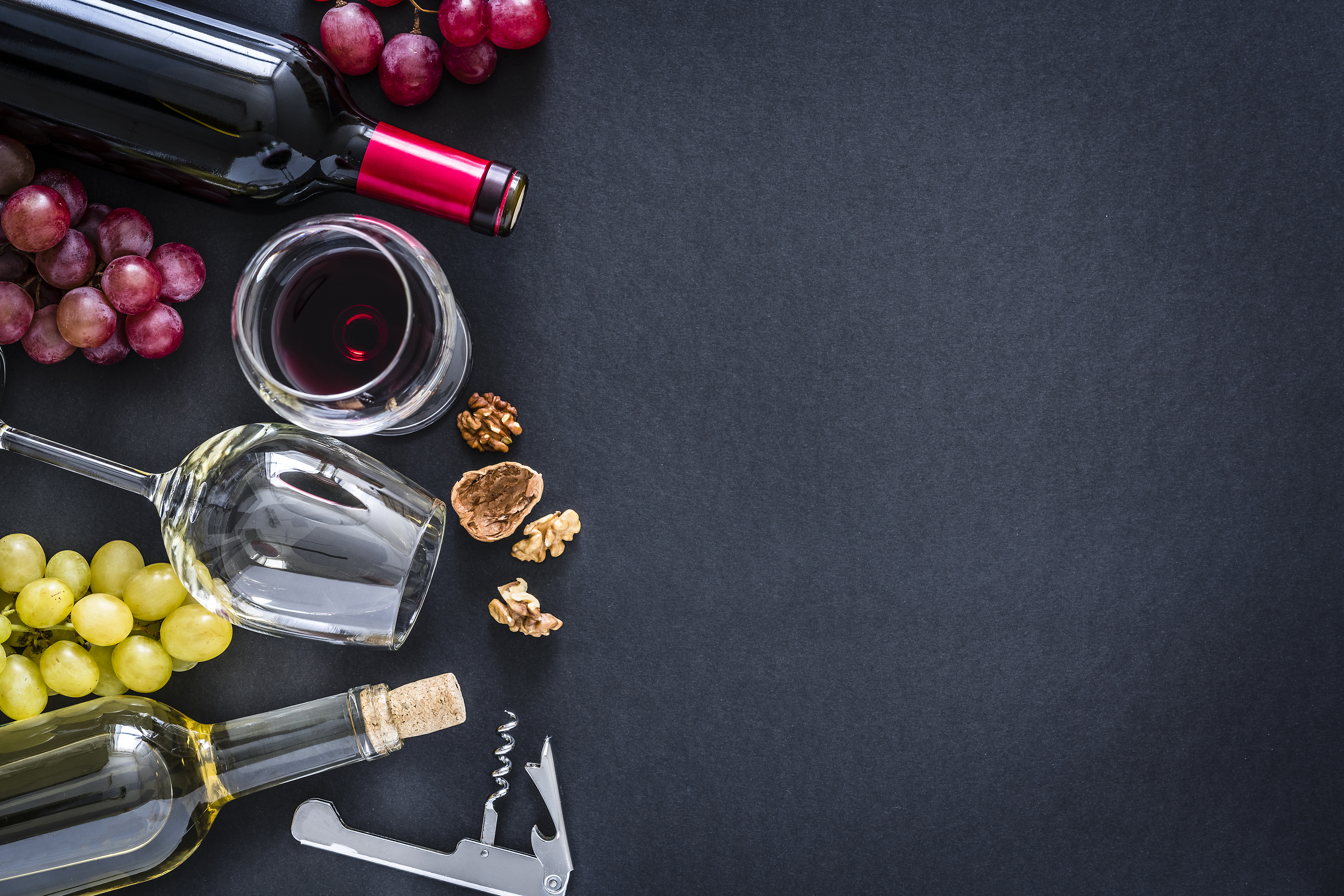 An overhead shot of wine bottles, stemware, grapes, and walnuts on a black backdrop 