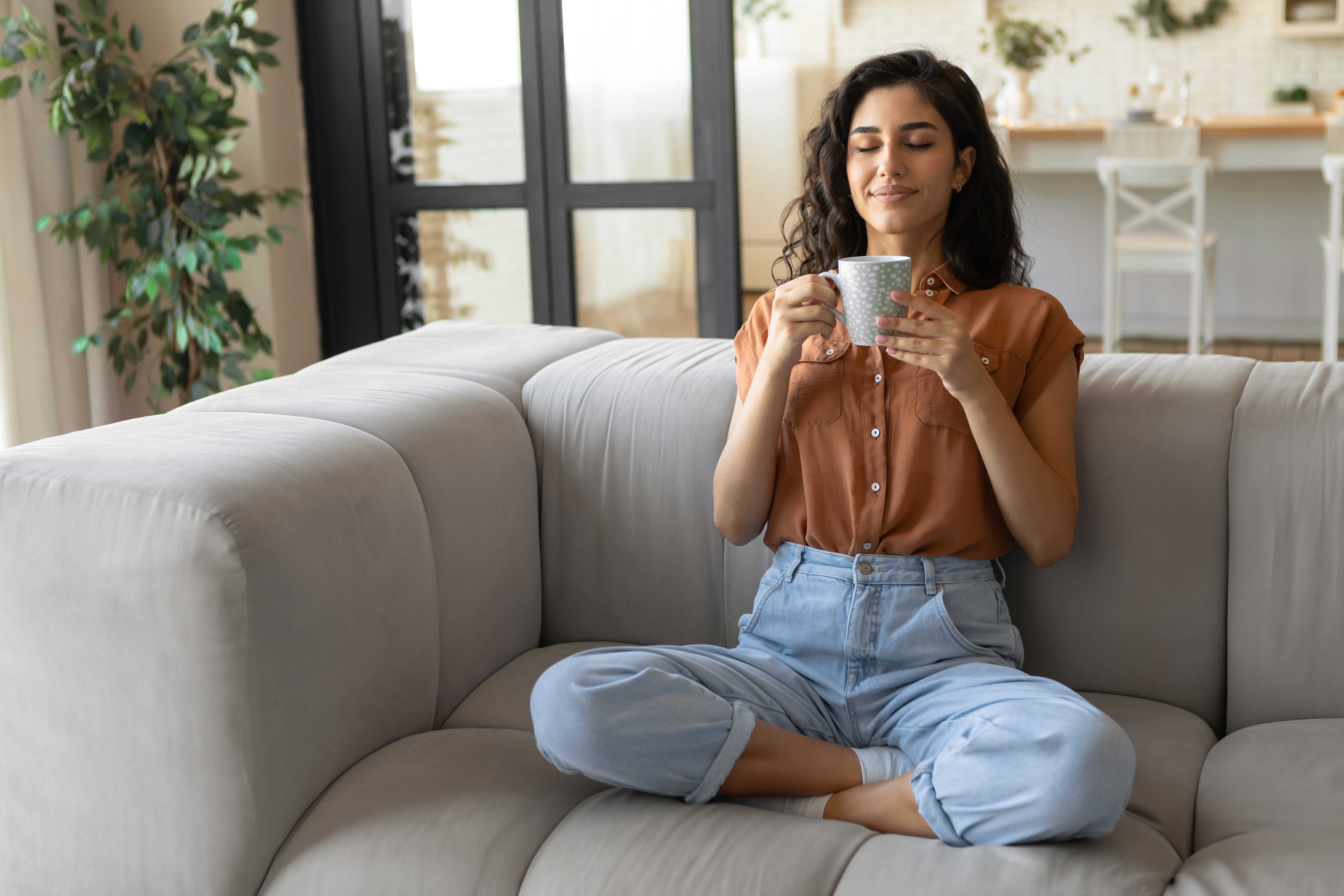 A young woman in a brown shirt sitting crisscross on a sofa, smiling, and holding a cup of tea