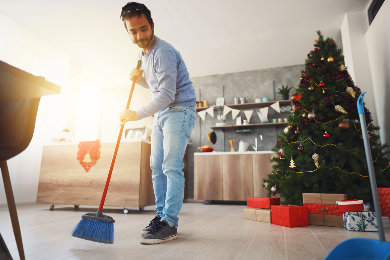 man sweeps his living room floor in front of his Christmas tree with gifts 