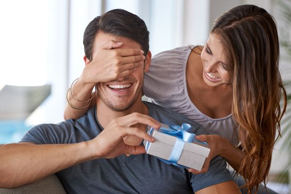 A woman covering a man’s eyes as she gives him a gift in a box 