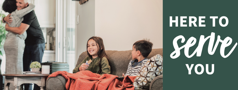 A young boy and girl smiling on a sofa while a man and woman hug in the background 