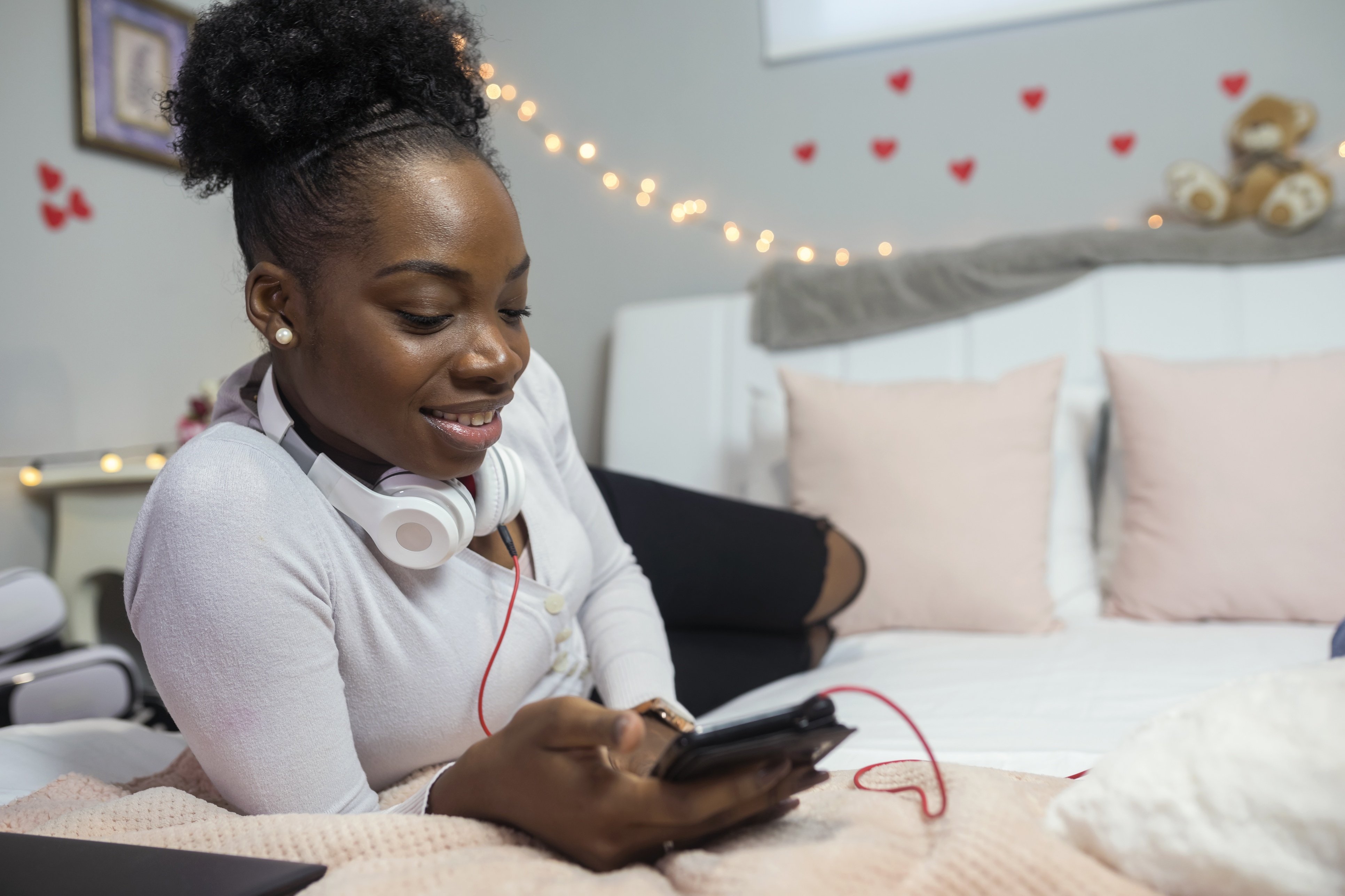 A teenage girl on a phone smiling while listening to music on a bed 