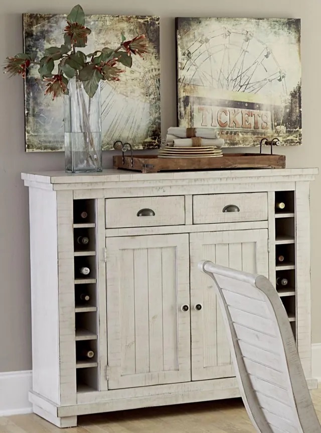 Photo of a white distressed wood wine cabinet with two storage drawers, two pullout doors, and wine bottle storage on each side with a vase of flowers and serving tray sitting on top. 