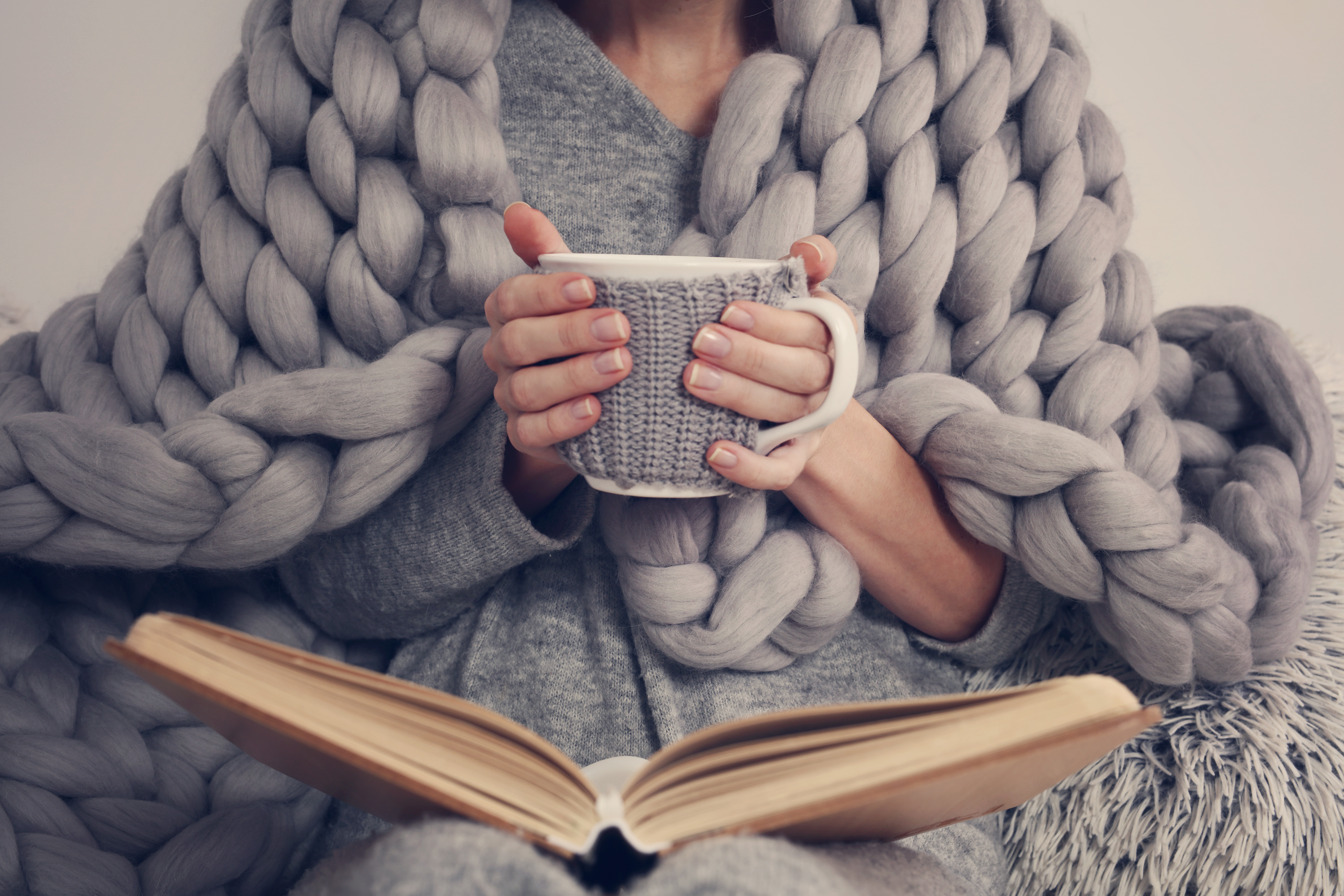 Woman wrapped in chunky-knit throw reading a book while enjoying a hot beverage 