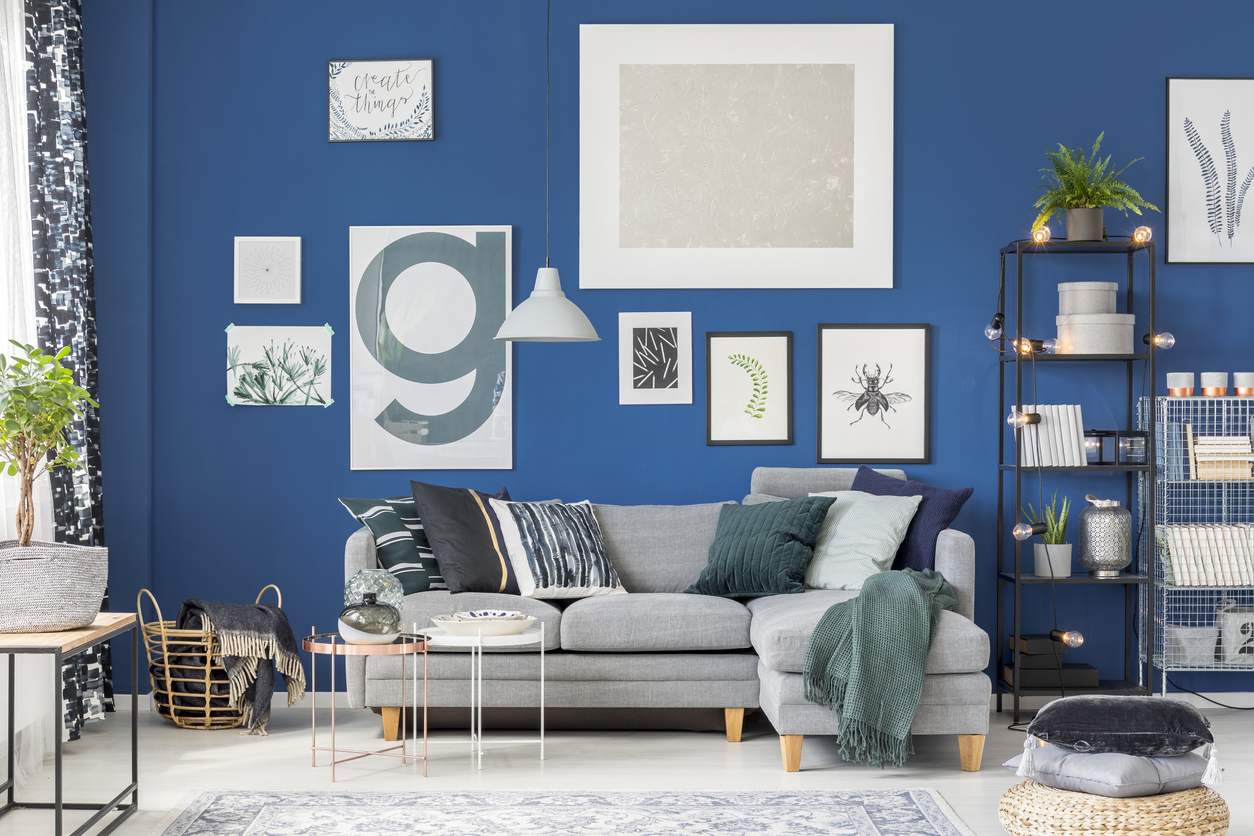pillows on pouf near grey sofa in cozy blue living room with posters, plants, and tables