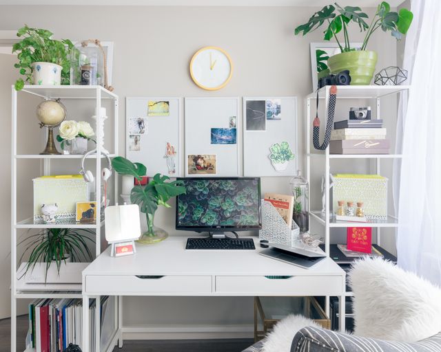 front view of a modern home office with white desk, white shelving on both sides, and a white fluffy chair. On the shelves is various decor, pants, and office supplies